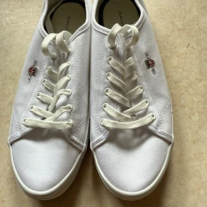 Vita sneakers från Gant i canvas - Snygga vita sneakers från Gant i klassisk modell med rund tå och platt sula. Skorna är i canvasmaterial och har snörning framtill. På sidan finns Gants broderade logga i rött, blått och vitt. Perfekta för en clean och stilren look.