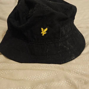 Svart bucket hat från Lyle & Scott - Svart bucket hat från Lyle & Scott med klassisk gul broderad örn-logga framtill. Hatten är tillverkad i 100% ekologisk bomull och har en avslappnad passform. Perfekt accessoar för att lyfta din streetwear-look.