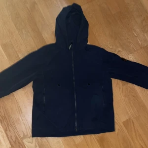 Mörkblå vindjacka från C.P. Company - Snygg mörkblå vindjacka från C.P. Company med huva och hel dragkedja framtill. Jackan har flera fickor, klassisk linsdetalj på ärmen och är tillverkad i lätt polyester. Går med på prut