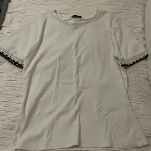 Vit t-shirt med spetsdetalj från ZARA - Vit t-shirt från ZARA i bomull med korta ärmar. Ärmsluten har en snygg virkad spetsdetalj i vitt och svart som ger en unik touch. Enkel och stilren modell med rund halsringning, perfekt för en clean look. Stl L men sitter som en S/M