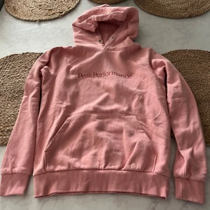Rosa hoodie från Peak Performance - Säljer en snygg rosa hoodie från Peak Performance med logga på bröstet. Tröjan har huva med snörningshål, magficka och ribbade muddar vid ärmslut och nederkant. Perfekt för en chill och avslappnad stil. Storlek:xs