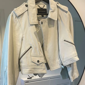 Vit skinnjacka från Banana Republic~M - Snygg vit skinnjacka från Banana Republic i bikerstil med coola dragkedjor och detaljer på axlarna. Jackan är tillverkad i äkta lammnappa och har klassisk krage, snedställda fickor och tryckknappar. Perfekt för dig som vill sticka ut med en stilren look.