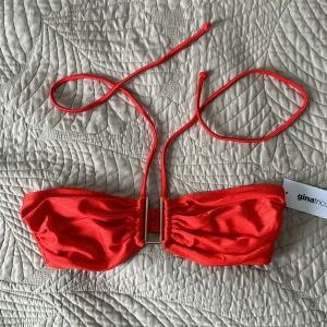 Röd bikiniöverdel från Gina Tricot - Snygg röd bikiniöverdel från Gina Tricot i modellen Alicia. Toppen har smala knytband i nacken och ett guldfärgat metallspänne framtill. Materialet är glansigt och stretchigt, perfekt för strandhäng. Färgen är intensivt röd och modellen är bandeau med rynkade detaljer. Ordinarie 229kr pris 100kr