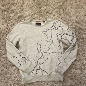 Ljusgrå sweatshirt från scotch&soda - Ljusgrå sweatshirt från Scotch & Soda med unikt svart kartmönster över hela tröjan. Tröjan har rund halsringning och långa ärmar. Tillverkad i mjuk bomull, perfekt för en chill och stilren look.