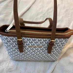 Michael Kors handväska med logomönster - Michael Kors handväska med klassiskt MK-logomönster i svart och vitt. Väskan har bruna handtag och detaljer i guld, samt dragkedja upptill. Insidan är fodrad med MK-tryck. Aldrig använt och är i nyskick! KÖPT I EGYPTEN