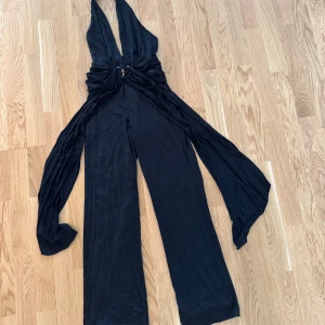 Svart byxdress från Oh Polly - Snygg svart byxdress från Oh Polly med djup v-ringning och vida ben. Byxdressen har knytband i midjan och är tillverkad i ett mjukt, följsamt material. Perfekt för dig som vill sticka ut med en trendig och elegant look.