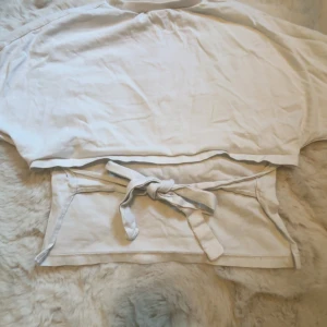 Vit croppad topp med knyt från Nelly - Säljer en vit croppad topp från Nelly i storlek S. Toppen har korta ärmar, rund halsringning och en snygg knytning i ryggen som ger en cool och avslappnad look. Perfekt att matcha med höga jeans eller kjol.