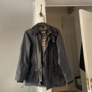 Svart Barbour bedale - Klassisk svart vaxad jacka från Barbour med rutigt innerfoder och krage i manchester. Jackan har stora fickor framtill, dragkedja och knappar. Perfekt för dig som gillar tidlös brittisk stil och vill ha en jacka som funkar hela hösten och våren.