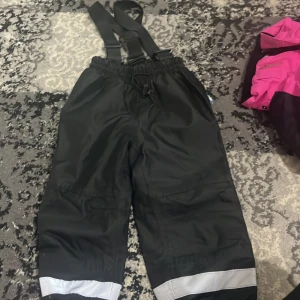 Svarta täckbyxor Peek-a-Boo 86/92 - Svarta täckbyxor från Peek-a-Boo i storlek 86/92. Byxorna har justerbara hängslen, resår i midjan och reflexdetaljer längst ner på benen. Perfekta för kalla dagar och lek utomhus.