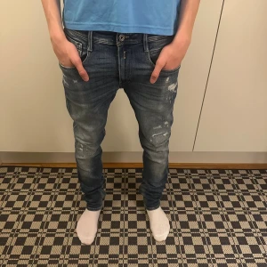 Replay anbass jeans - Säljer ett par blå jeans med snygga slitningar och tvättade detaljer. Jeansen har tapered passform, fem fickor och klassisk dragkedja. Materialet är jeans och färgen är mörkblå med ljusare partier. Perfekta för en avslappnad och trendig look.