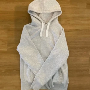 Russemerch hoodie  - En stilren ljusgrå hoodie med huva och vita snören. Tröjan har lång ärm, känguruficka framtill och ribbade muddar vid ärmslut och nederkant. Perfekt för en avslappnad look.