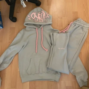 Grå hoodie med rosa detaljer från SynaWorld  - Säljer en ljusgrå hoodie från SynaWorld med rosa snören och coolt rosa tryck på huvan. Hoodien har känguruficka och mjukt material, perfekt för chill dagar. Matchande byxor med rosa snörning och tryck ingår.