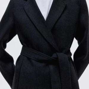 Svart lång kappa med bälte - Stilren svart kappa i klassisk modell med knytbälte i midjan och långa ärmar. Kappan har breda slag, rak passform och är tillverkad i ett mjukt ullmaterial. Perfekt för dig som vill ha en tidlös och snygg look under höst och vinter.