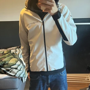 Vit vindjacka från Sail Racing  - Säljer denna vita vindjackan från Sail Racing med svarta detaljer och dragkedja framtill. Jackan har hög krage, lång ärm och logga på bröstet och ärmen. Tillverkad i polyester, perfekt för blåsiga dagar. Använd max 2-3 gånger. Nypris 2000kr.