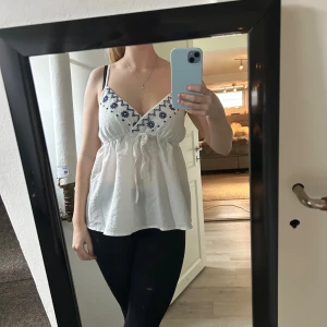 Vit broderad linnetopp med knyt - Säljer en söt vit linnetopp med blå broderade blommor vid bysten och knyt under bysten. Toppen har v-ringning, tunna axelband och en luftig passform. Perfekt för varma dagar och ger en fräsch sommarkänsla. Strl: M, passar S🩷