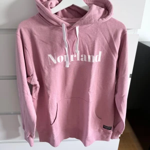 Rosa hoodie från SORTN COMPANY - Säljer en rosa hoodie från SORTN COMPANY med vit text 'Norrland' på bröstet. Tröjan har huva med vita snören, känguruficka och är tillverkad i mjuk bomull. Perfekt för dig som gillar streetwear och vill ha en chill look.