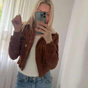 Brun manchester bomberjacka med fuskpäls - Säljer en brun bomberjacka i manchester med croppad passform och mysig fuskpäls på kragen. Jackan har dragkedja framtill, ribbade muddar och coola detaljer på ärmarna. Perfekt för dig som vill ha en trendig och chill look.
