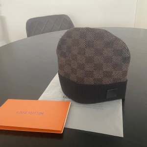Svart Louis Vuitton mössa - Exklusiv mössa från Louis Vuitton i brunt med klassiskt rutigt Damier-mönster. Mössan har en ribbad kant och en diskret LV-logga framtill. Tillverkad i mjukt material som känns lyxigt och passar perfekt för att lyfta din vinterstil.