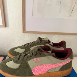 Puma Palermo sneakers oliv/rosa - Säljer ett par Puma Palermo sneakers i olivgrönt och beige tyg med detaljer i mocka. Skorna har en rosa Puma-logga på sidan, vinröd insida och klassisk gummisula. Snygg retrostil med snörning och rund tå, perfekta för dig som vill sticka ut lite extra.