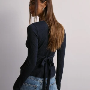 Mörkblå omlottopp med knyt - Supersnygg mörkblå omlottopp med lång ärm och v-ringning. Toppen har ribbad struktur och knyt i midjan som ger en smickrande siluett. Perfekt att styla med jeans för en trendig look.