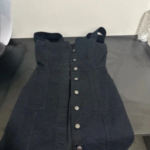 Svart jeansklänning med knappar - Snygg svart jeansklänning med breda axelband och knappar hela vägen framtill. Klänningen har två stora fickor framtill och en figurnära passform som ger en cool och avslappnad look.