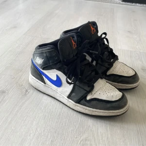 Nike Air Jordan 1 Mid svart/blå 38.5 - Nike Air Jordan 1 Mid i svart, vitt och blått med klassisk Jordan-logga på plösen i orange. Skorna har snörning, rund tå och platt sula. Swoosh-loggan är blå och materialet är skinn och syntet. Perfekta för dig som gillar streetstyle och sneakers med attityd.