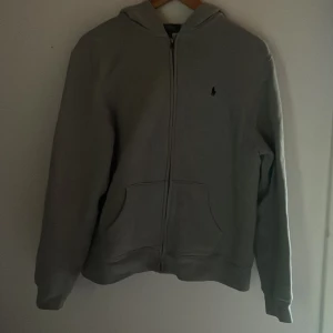 Grå ZipHoodie från Polo Ralph Lauren  - Klassisk grå hoodie från Polo Ralph Lauren i storlek 163-174. Tröjan har dragkedja framtill, huva och två stora fickor. Liten broderad logga på bröstet. Perfekt för chill dagar och enkel att matcha med jeans eller joggers.