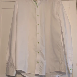 Vit skjorta Dressmann L med gröna knappar - Vit skjorta från Dressmann i storlek L med snygga ljusgröna knappar. Skjortan är tillverkad i en easy care bomullsblandning och har klassisk krage samt långa ärmar. Perfekt för dig som gillar stilrena plagg med en liten twist.