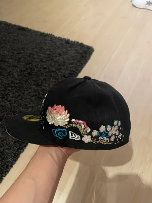 Svart New Era keps med blommor - Säljer en svart New Era 59FIFTY keps med broderade blommor i rosa, guld och blått på sidan. Framtill finns ett vitt SF-logo och detaljer i vitt. Skärmen är platt och undersidan är vit. Perfekt för dig som vill sticka ut med snygga detaljer. kepsen är 7 1/8 men jag skulle säga att den passar som en 7 1/4 också. 