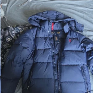 Mörkblå pufferjacka Polo Ralph Lauren - Säljer en mörkblå pufferjacka från Polo Ralph Lauren med huva och dragkedja. Jackan är typ helt ny och aldrig används utomhus