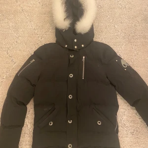 Svart dunjacka Moose Knuckles - Svart dunjacka från Moose Knuckles med vit päls på huvan, silverfärgade detaljer och dragkedjor på ärmarna. Jackan har broderad logga på insidan och ikonisk metallpatch på ärmen. Flera fickor med knappar och dragkedja framtill. Perfekt för kalla dagar.