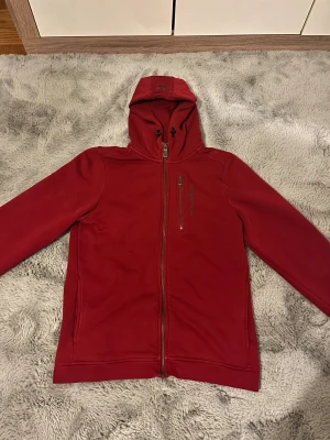 Röd fleecejacka Sail Racing med huva - Röd fleecejacka från Sail Racing med huva och hel dragkedja framtill. Jackan har en bröstficka med dragkedja och snygga detaljer vid snörningen på huvan. Materialet är mjuk fleece med inslag av bomull och polyester, perfekt för kyliga dagar.