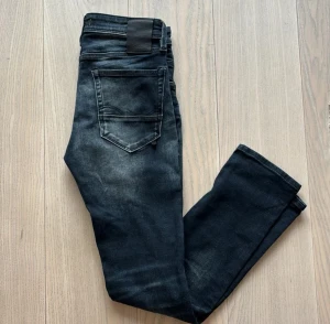 Svarta Jack & Jones jeans med slitningar - Snygga svarta Jack & Jones jeans med coola slitningar och tvättade detaljer. Klassisk femficksmodell med raka ben och normal passform. Jeansen har en mörk tvätt med ljusare partier och är tillverkade i mjukt denimtyg som sitter skönt hela dagen.