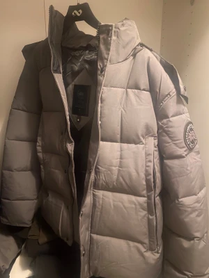 Grå Canada Goose  - Säljer en grå pufferjacka från Canadian Goose med patch på ärmen och hög krage. Jackan har dragkedja och tryckknappar framtill, samt två sidofickor. Den är quiltad och har en oversized look, perfekt för kalla dagar.
