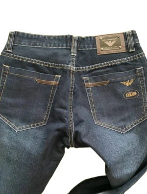 Mörkblå jeans från Armani - Snygga mörkblå jeans från Armani med klassisk femficksdesign, kontrastsömmar och broderad logga på bakfickan. Jeansen har metallmärke bak i midjan och är tillverkade i slitstarkt denim. Perfekta för dig som gillar stilrena och exklusiva jeans.