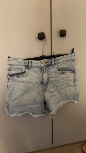 Ljusblå jeansshorts med fransad kant - Snygga ljusblå jeansshorts med klassisk femficksdesign och fransad kant nedtill. Shortsen har knapp och dragkedja framtill samt bälteshällor. Perfekta för varma dagar och har en chill vibe med sin slitna look.