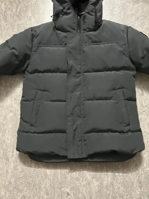 Svart pufferjacka från Canada Goose - Säljer en svart pufferjacka från Canada Goose i storlek M. Jackan har en stilren look med broderad logotyp på ärmen, snörning i huvan och två sidofickor. Materialet är syntet och polyester, perfekt för kalla dagar. Dragkedja framtill med täckande vindslå.