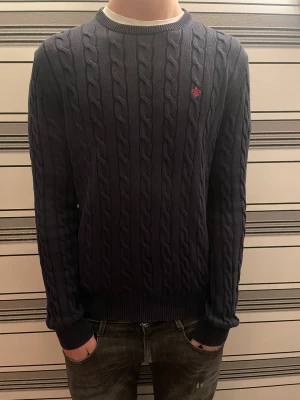 Mörkblå kabelstickad tröja Morris - Snygg mörkblå kabelstickad tröja från Morris med rund hals och diskret röd logga broderad på bröstet. Tröjan har ribbade muddar vid ärmslut och nederkant, och är långärmad. Perfekt för dig som gillar klassisk stil med en modern touch.