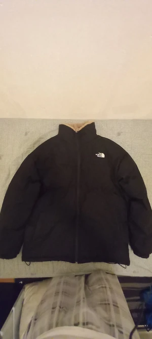 Svart teddyjacka från The North Face - Mysig svart teddyjacka från The North Face med beige fluffigt foder på insidan. Jackan har hög krage, dragkedja framtill och logga på bröstet. Perfekt för kalla dagar när du vill vara både varm och stilren.