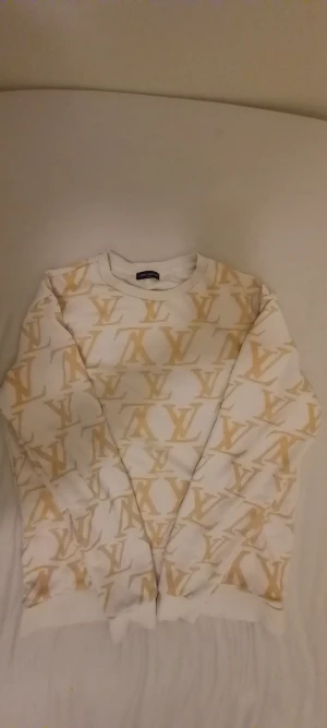 Vit och guld Louis Vuitton sweatshirt - Exklusiv sweatshirt från Louis Vuitton i vitt med gul LV-logga över hela tröjan. Rund hals, långärmad och tillverkad i mjuk bomull. Snyggt mönster som sticker ut och ger en lyxig vibe. Made in Italy, perfekt för dig som gillar designer och streetstyle.