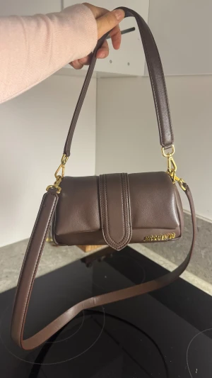 Brun axelväska från Jacquemus  - Säljer en brun axelväska från Jacquemus i äkta skinn med guldiga metalldetaljer och avtagbar axelrem. Väskan har en stilren, avlång form och stängs med lock. Jacquemus-logga i guld framtill. Dustbag medföljer.