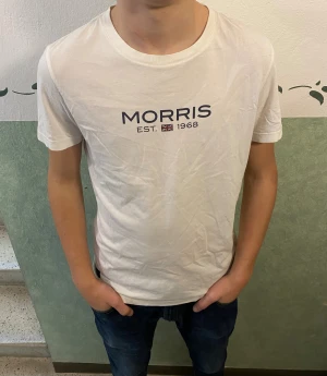 Vit t-shirt från Morris med tryck - Vit t-shirt från Morris med svart tryck på bröstet där det står 'MORRIS EST. 1968' och en liten brittisk flagga. Klassisk rund hals och korta ärmar. Skön och mjuk i bomull, perfekt till jeans eller shorts.