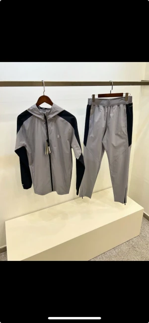 Grå träningsset från Under Armour - Snyggt tvådelat träningsset från Under Armour i ljusgrått med svarta detaljer. Setet består av en zip-hoodie med huva och matchande byxor med dragkedja vid benslut. Perfekt för dig som vill ha en sportig och clean look.