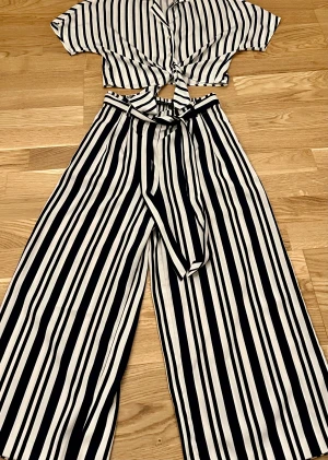 Randig tvådelad kostym med knyt - Tvådelad kostym från Bershka med marinblå och vita ränder. Croppad skjorta med knyt framtill, kort ärm och krage. Vida byxor med hög midja och matchande bälte. Lätt och luftig 