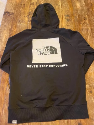 Svart hoodie från The North Face - Svart hoodie från The North Face med stor logga och texten 'Never Stop Exploring' på ryggen. Klassisk huva, ribbade muddar och magficka framtill. Materialet är mjuk bomull och polyester. Snygg och enkel design med vit logga även på bröstet.