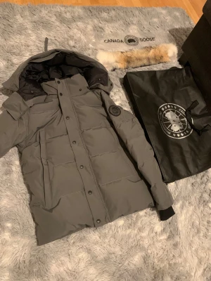Grå dunjacka från Canada Goose -  Kom med bud. Säljer en grå dunjacka från Canada Goose med avtagbar päls på huvan. Jackan har svart logotyp på ärmen, dragkedja och knappar framtill samt ribbade muddar. Perfekt för kalla vinterdagar och har en clean, modern look.