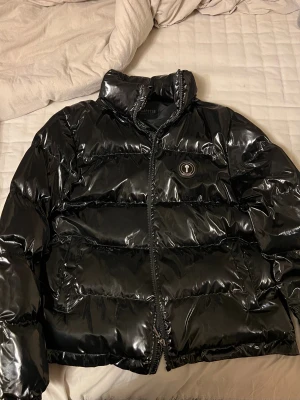 Svart pufferjacka från Trapstar M - Snygg svart pufferjacka från Trapstar med glansig finish och broderad logga på ryggen. Har bara använt den en vinter och bara några gånger. Jackan har hög krage, dragkedja framtill och rund patch med 'It's a Secret' på bröstet. Perfekt för kalla dagar och streetwear-stil. 