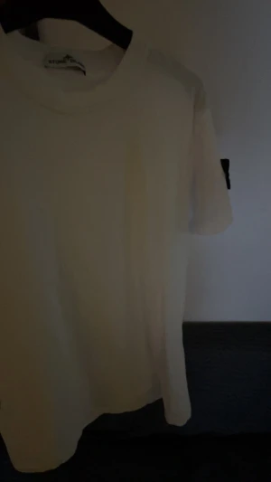 Vit t-shirt från Stone Island - Vit t-shirt från Stone Island med klassisk rund hals och korta ärmar. På vänster ärm sitter den ikoniska svarta patchen med gul broderad logga. T-shirten är gjord i mjuk bomull och har en clean, stilren look som funkar till streetwear.