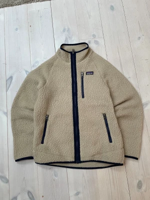 Beige fleece från Patagonia - Mysig beige fleece från Patagonia i storlek L barn. Jackan har hel dragkedja framtill, tre fickor med dragkedja och blått kantband. Tillverkad i 100% återvunnen polyester med fluffig yta och hög krage. Perfekt för kyliga dagar och outdoor-häng.