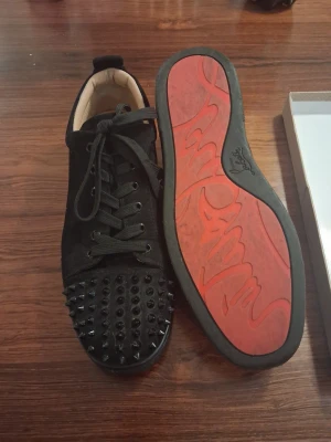Christian Louboutin svarta sneakers med nitar - Svarta sneakers från Christian Louboutin med ikonisk röd sula och svarta nitar på tån. Skorna har snörning och är tillverkade i mocka med skinnfoder. Medföljer original dustbag och låda. Perfekt för dig som vill sticka ut med lyxig streetstyle. Storlek 44 men passar 43 också. Priset är ej hugget i sten